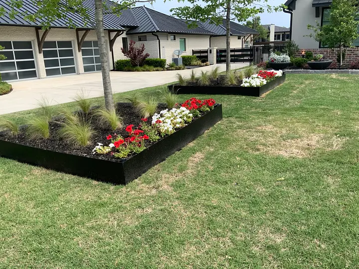 Flower beds with red and white blooms and grasses in front of a modern house with a metal roof and large garage.