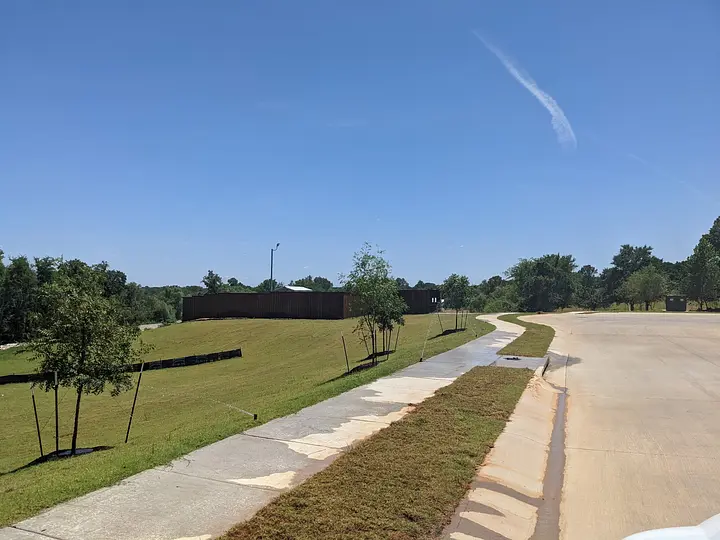 Curved sidewalk beside a grassy slope and trees under a clear blue sky with a single cloud streak.